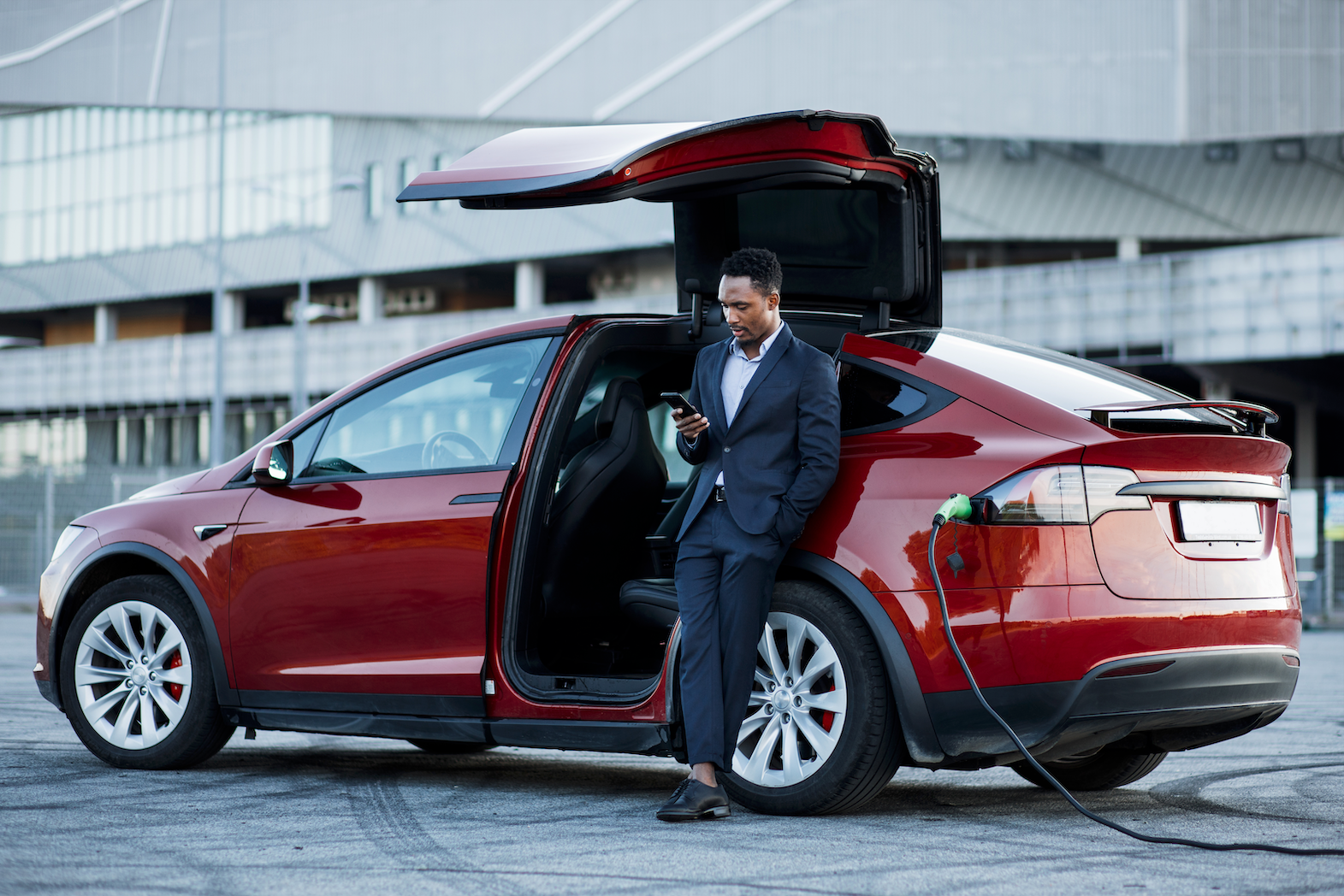 Man in a suit looking at his phone, leisurely leaning against his car while charging his electric vehicle