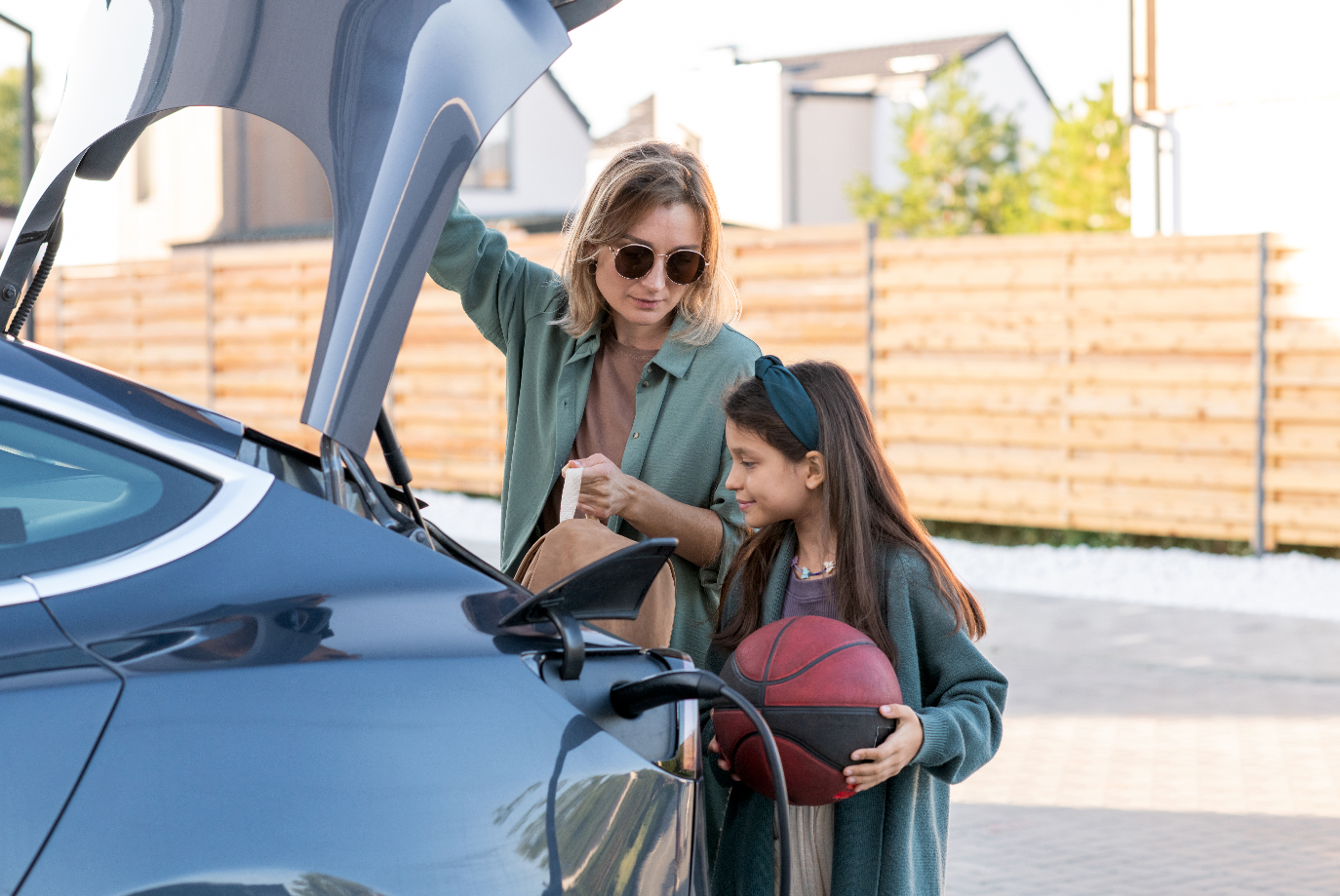 A lady opening the trunk of an electric vehicle, next to a child holding a basketball and smiling